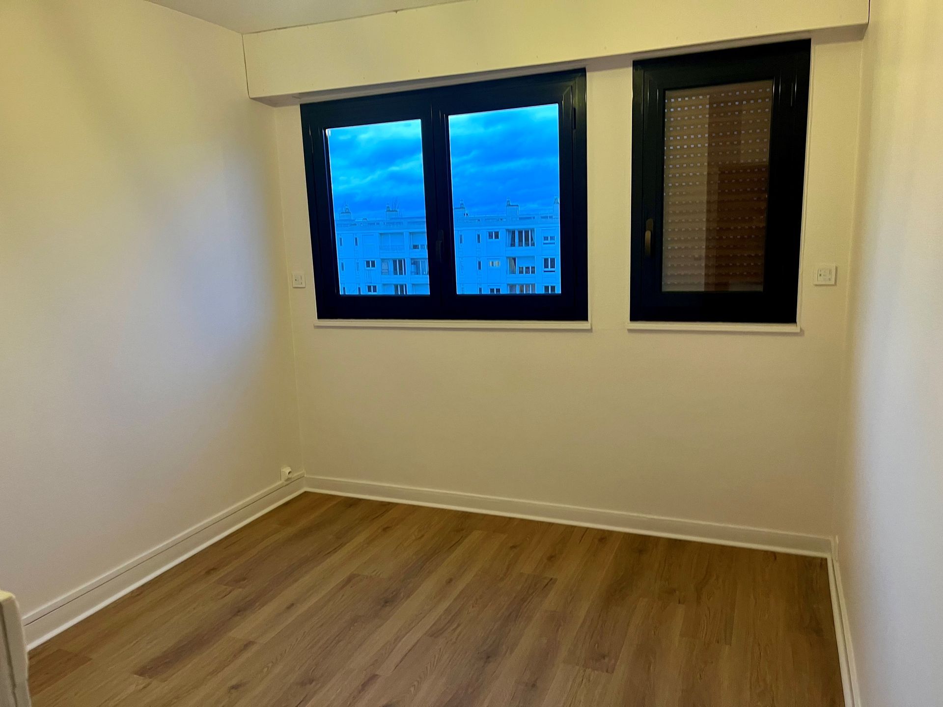 Une chambre vide aux murs clairs, au parquet en bois et dotée de deux fenêtres à cadre sombre donnant sur un ciel bleu.