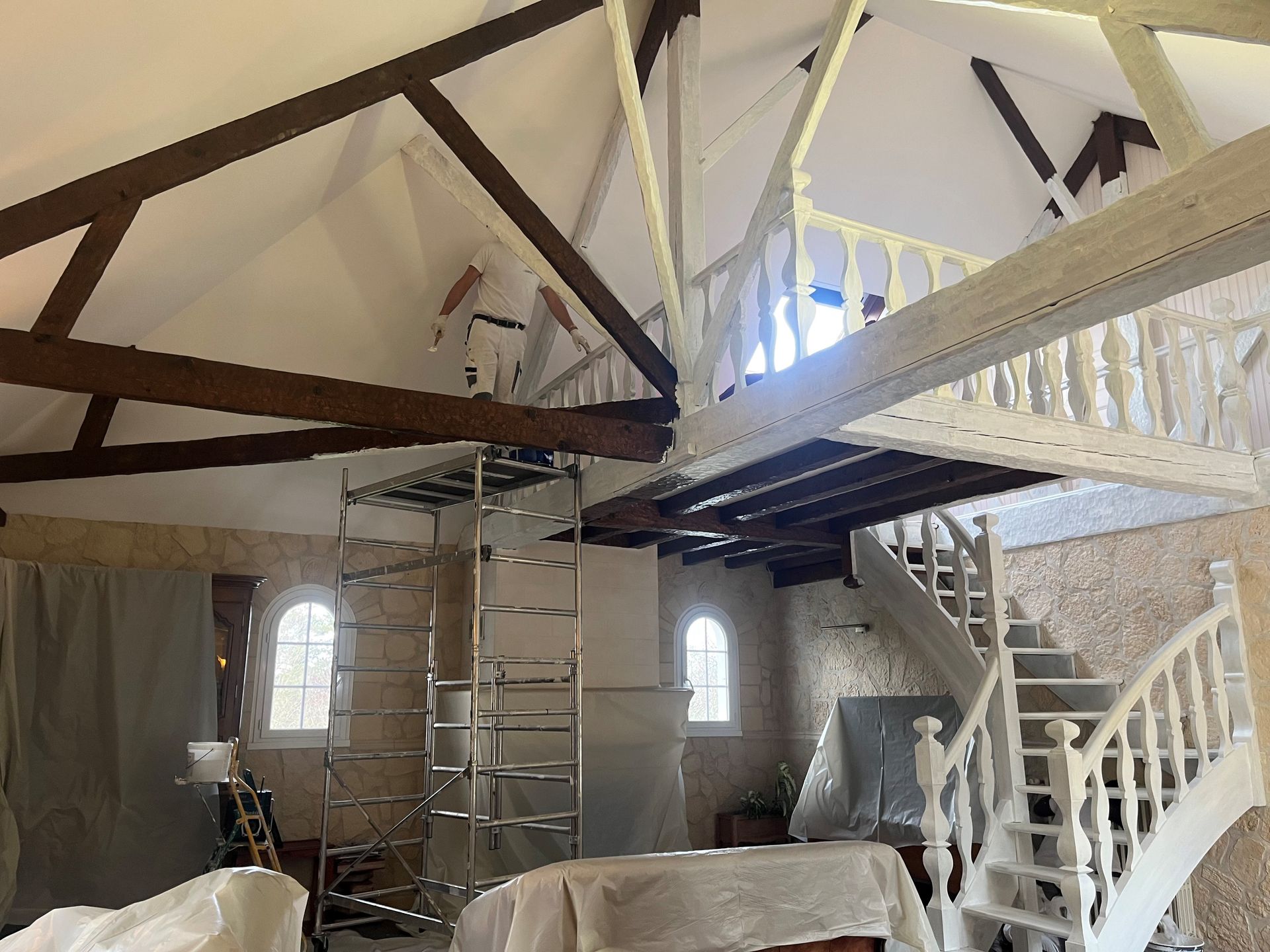 Une personne, perchée sur un échafaudage, peint les hautes poutres d'une pièce rustique aux murs de pierre apparente, avec un escalier et une mezzanine.