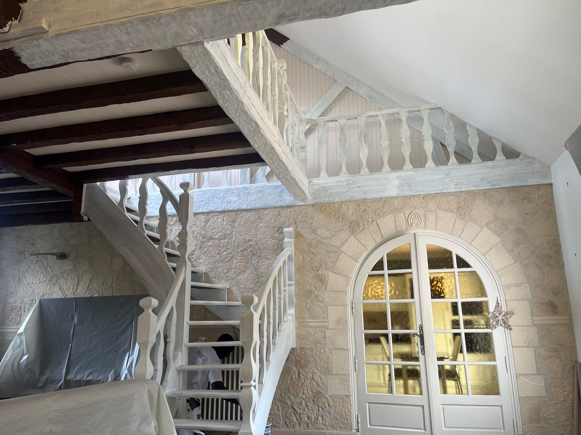 Un intérieur rustique avec une porte cintrée encadrée de pierre, un escalier blanc et une balustrade de mezzanine sous des poutres apparentes.