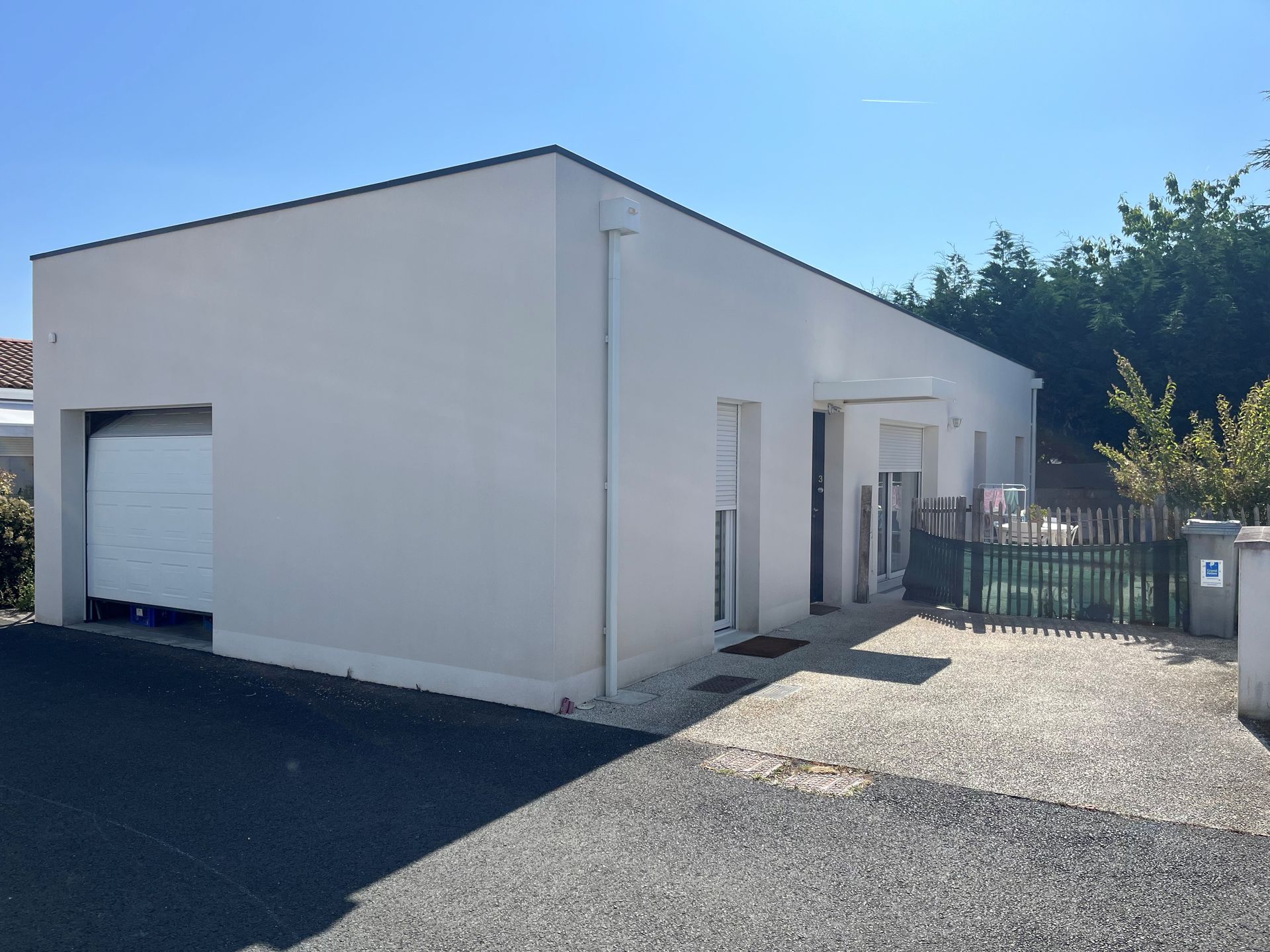 Une maison moderne de plain-pied, de couleur claire, avec un toit plat, une porte de garage blanche et une petite cour clôturée.