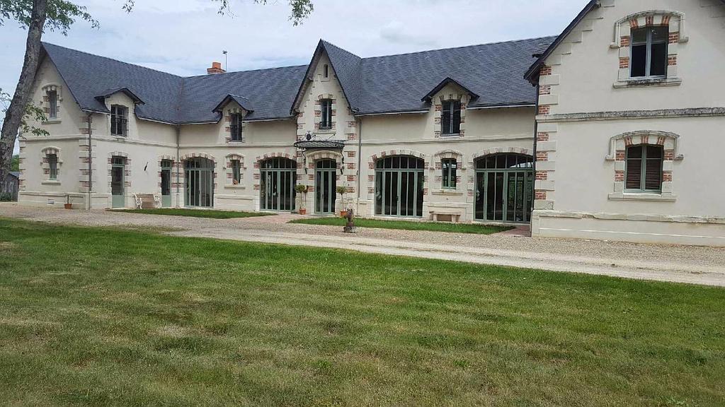 Une grande maison de campagne couleur crème, en forme de L, avec un toit à pignon sombre, de nombreuses portes vitrées et une pelouse au premier plan.