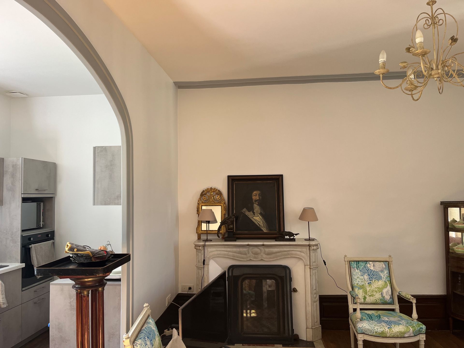 Dans une pièce ornée d'un lustre décoratif, d'un fauteuil à motifs floraux et d'une arche, se trouve une cheminée richement décorée avec un tableau encadré.