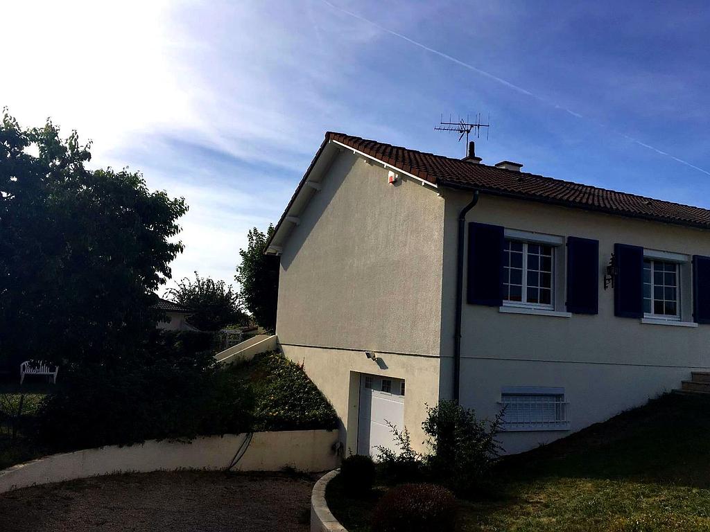 Une maison couleur crème, aux volets foncés et au toit de tuiles, vue depuis une allée de gravier légèrement surélevée sous un ciel bleu.