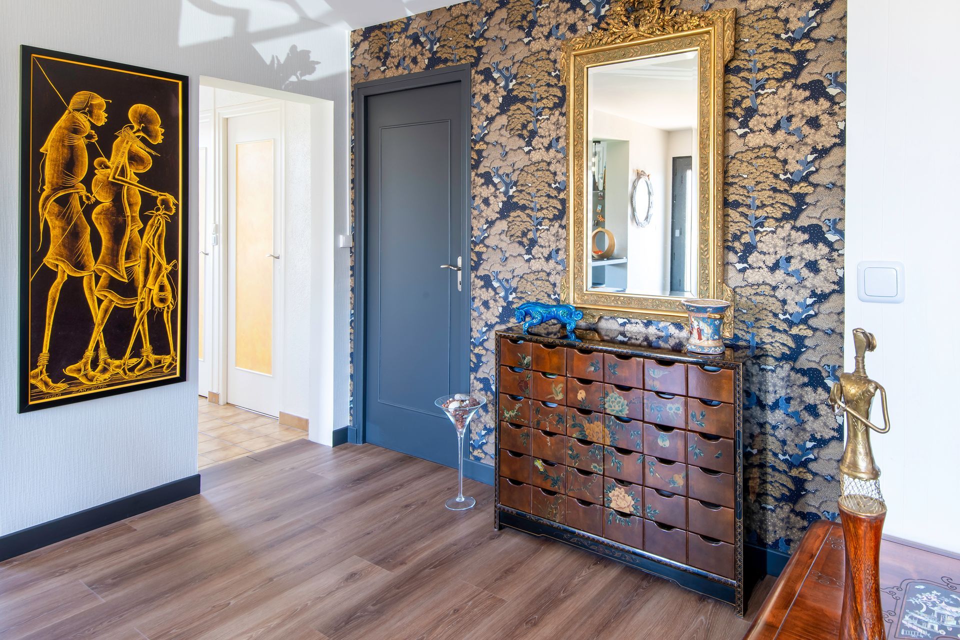 Un couloir avec une porte bleu foncé, du papier peint à motifs, un miroir à cadre doré, une commode à plusieurs tiroirs et des décorations murales.