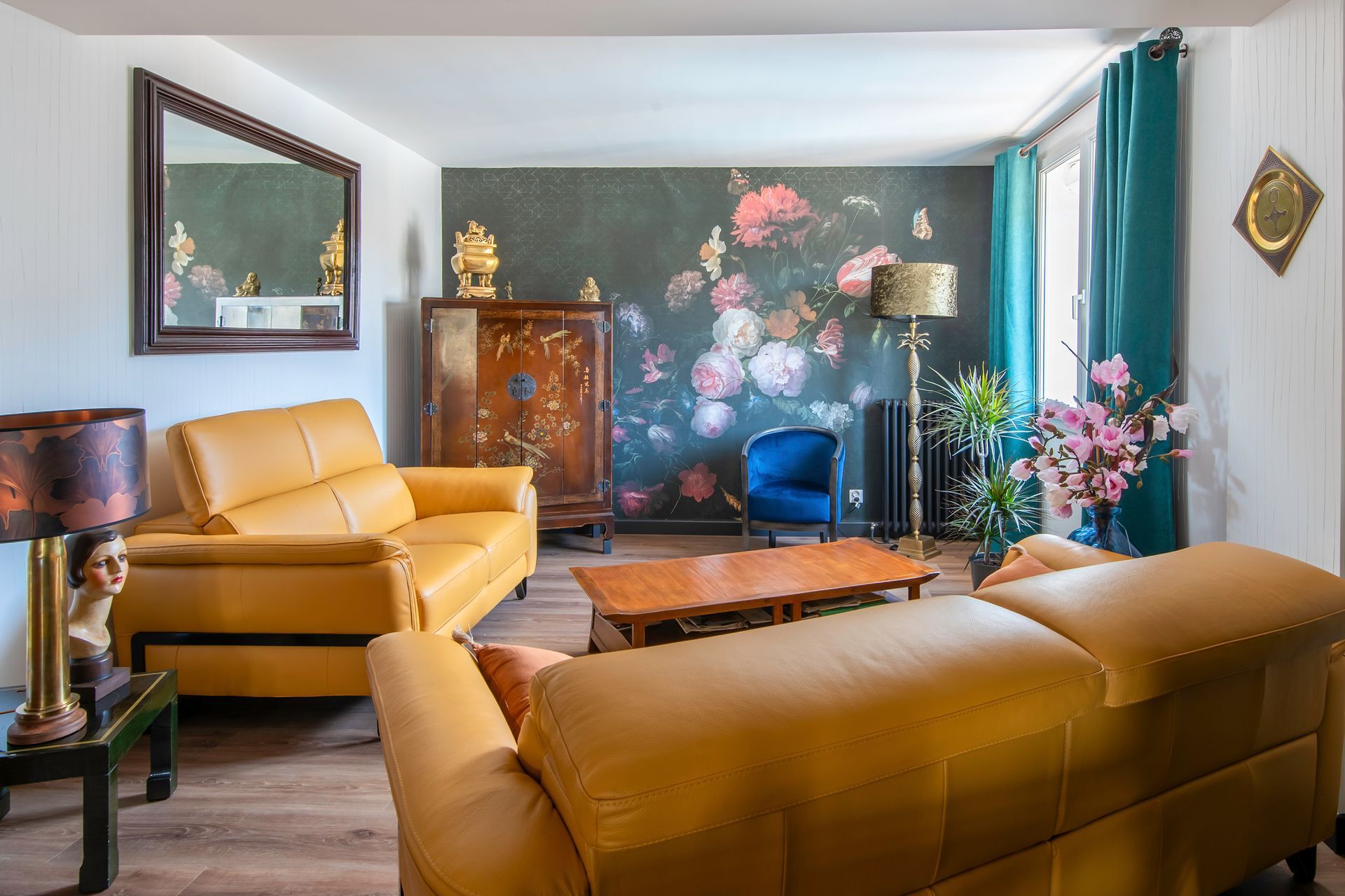 Le salon comprend deux canapés en cuir jaune, une table basse en bois, un fauteuil en velours bleu et du papier peint à motifs floraux.