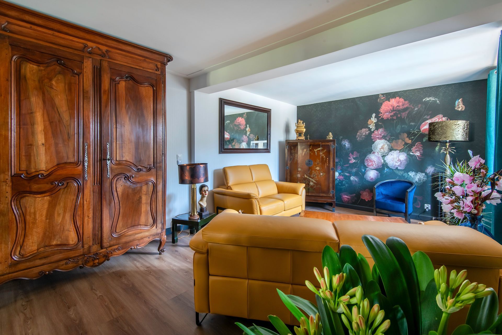 Le salon comprend une grande armoire en bois, des sièges jaunes, un mur d'accent floral foncé et une plante au premier plan.