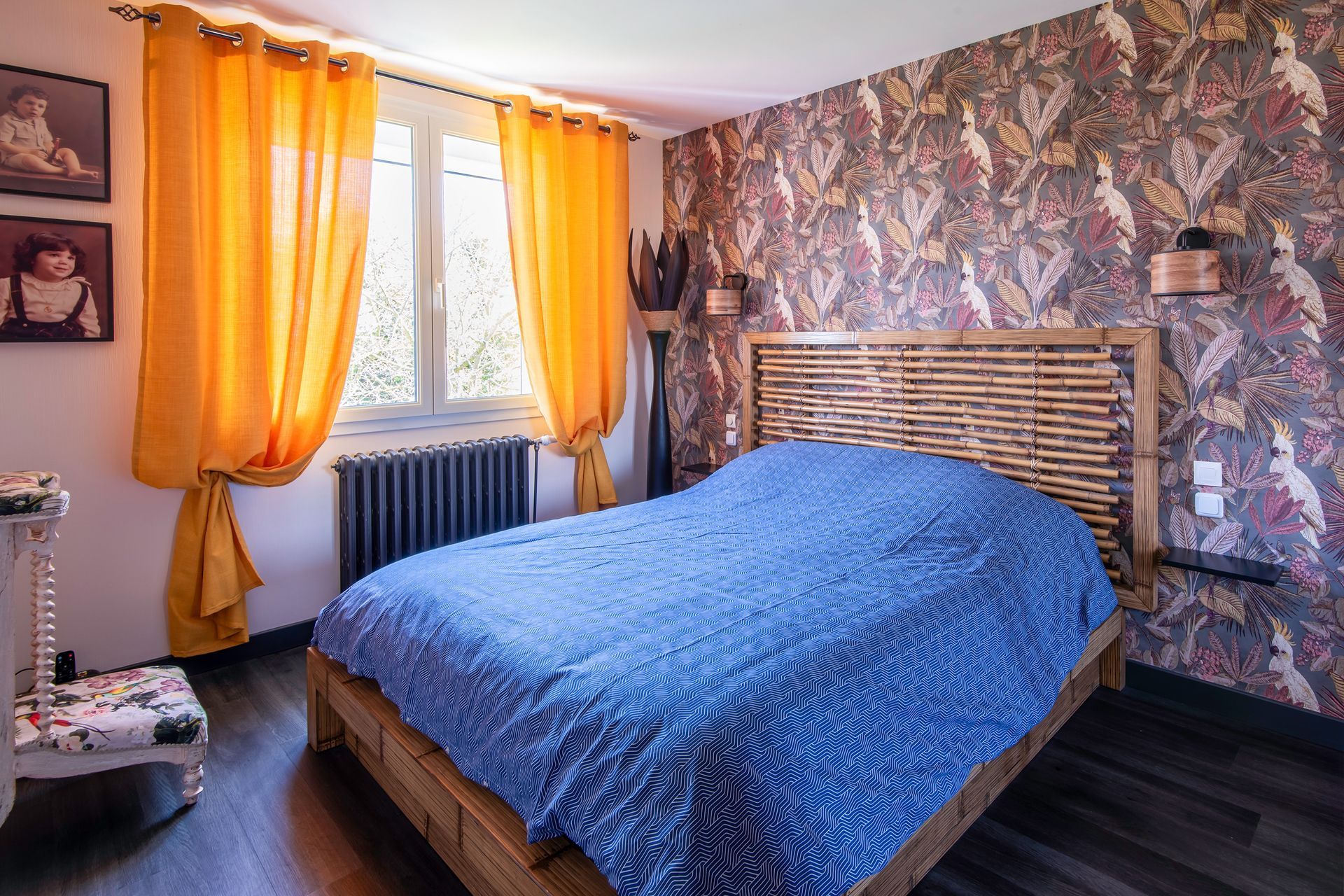 Une chambre avec une couette bleue sur un lit en bois, du papier peint à motifs, des rideaux jaunes et une fenêtre au-dessus d'un radiateur.
