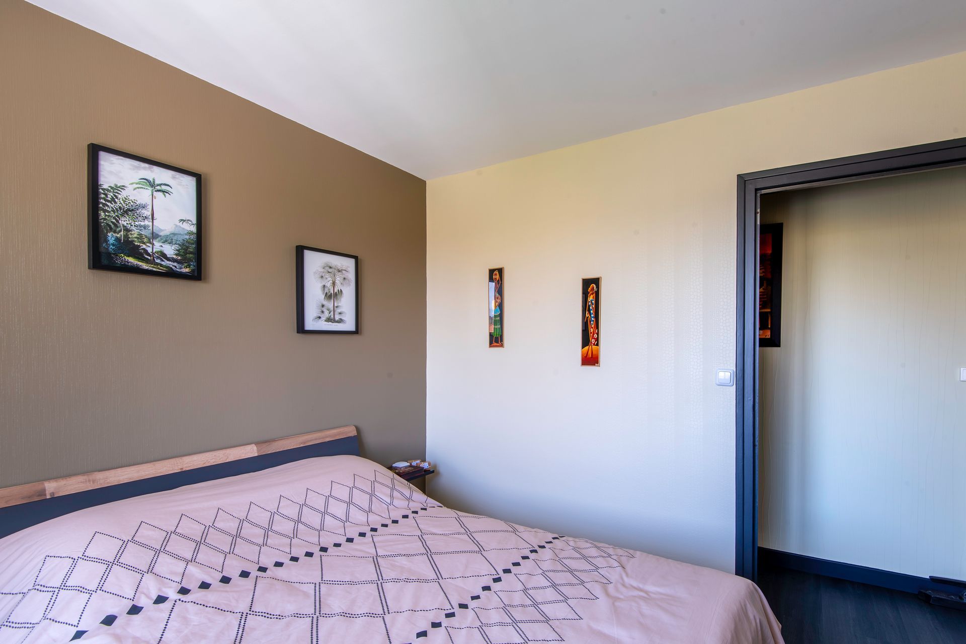 Un lit recouvert d'une couette rose à motifs dans une chambre aux murs taupe et crème ornés de tableaux encadrés.