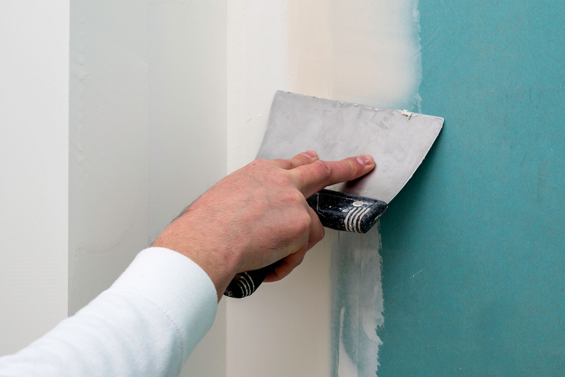 Une main utilise un couteau à mastic métallique pour appliquer un enduit blanc sur un joint d'un mur bleu turquoise.