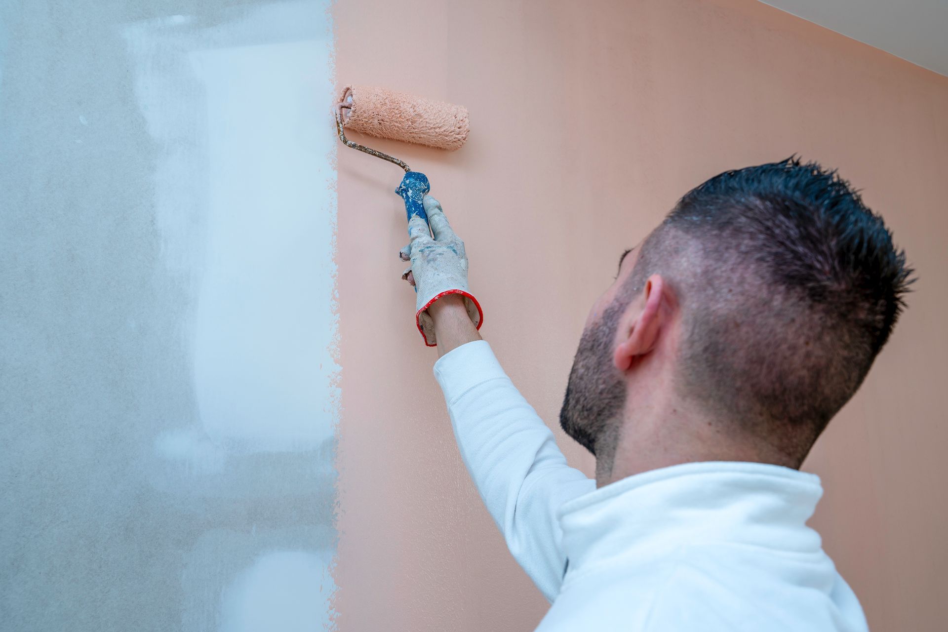 Une personne portant une chemise blanche et un gant peint un mur avec un rouleau couleur pêche.