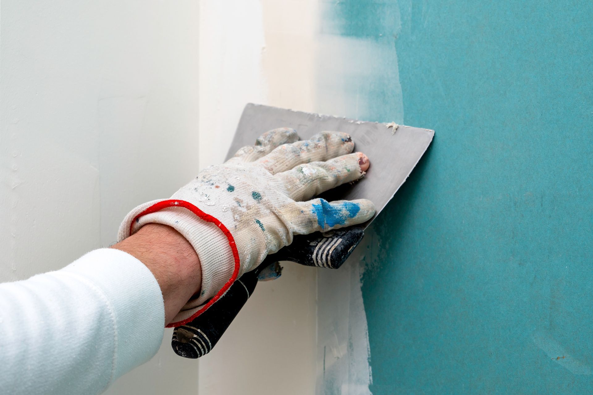 Une personne portant un gant taché de peinture utilise un couteau à mastic métallique pour appliquer un enduit à joints blanc sur un joint de cloison sèche turquoise.