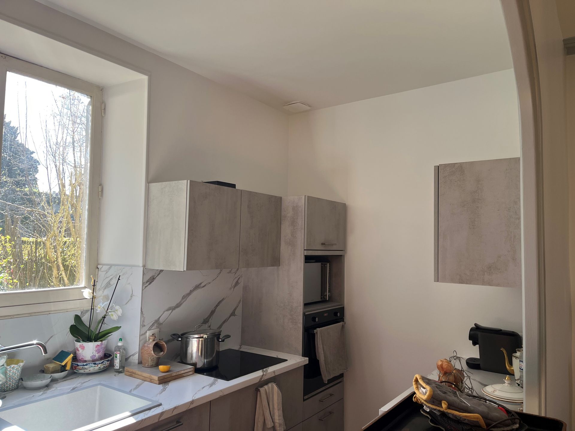 Un coin cuisine lumineux avec un évier blanc, des plans de travail clairs, des meubles gris et une fenêtre laissant entrer la lumière naturelle.