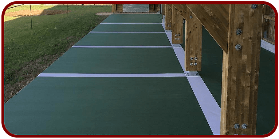 Un porche ou une terrasse avec une surface verte, divisée par des bandes blanches, et comportant des poteaux de soutien en bois sur un côté.