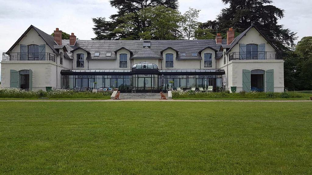 Un grand manoir de campagne à plusieurs étages, avec un rez-de-chaussée vitré, des lucarnes et une vaste pelouse au premier plan.