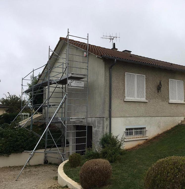 L'extérieur d'une maison de couleur claire, avec des échafaudages métalliques installés contre un mur latéral pour des travaux de construction ou de rénovation.