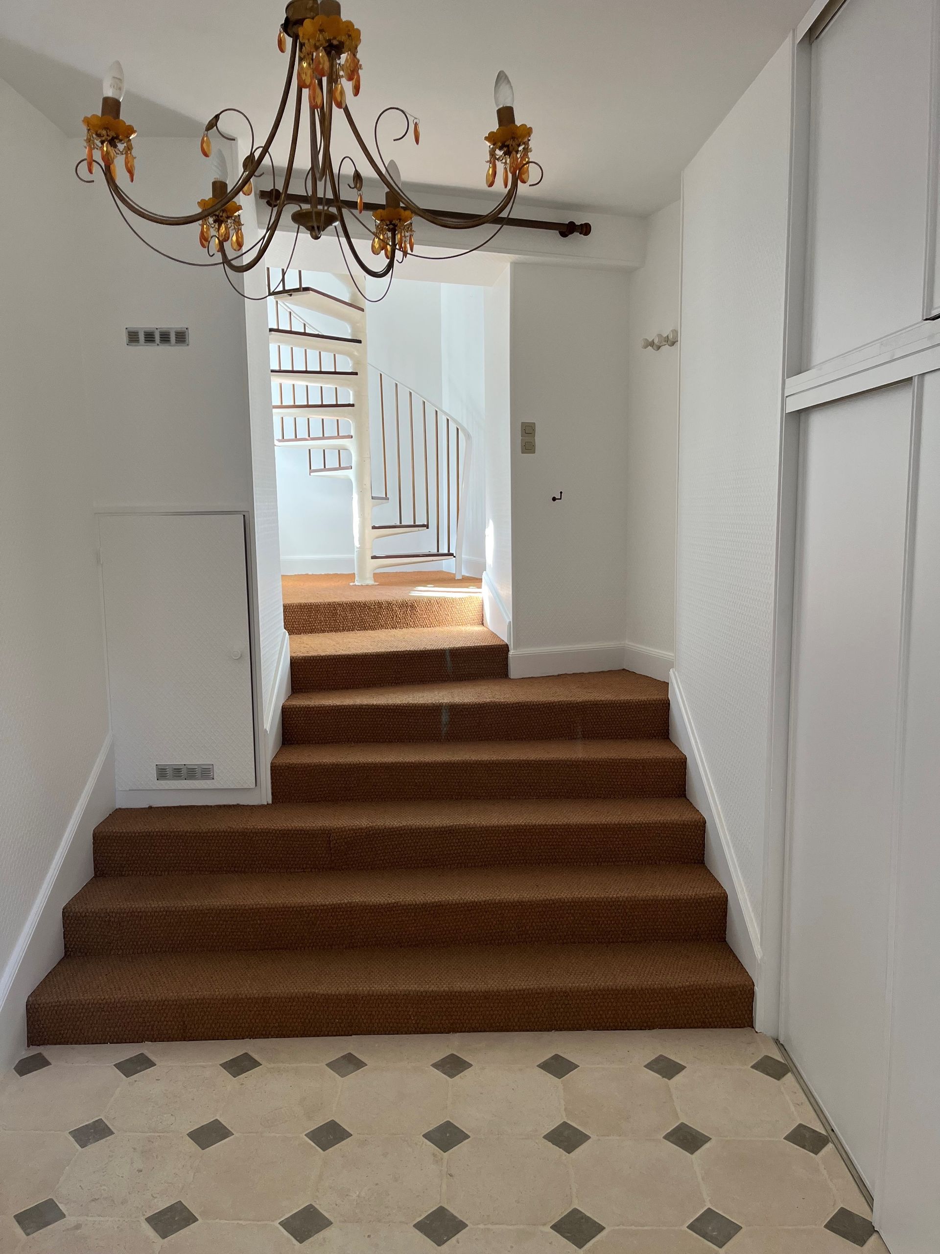 Un escalier recouvert de moquette marron mène à un palier où se trouve un escalier en colimaçon blanc, sous un lustre, dans un hall blanc.