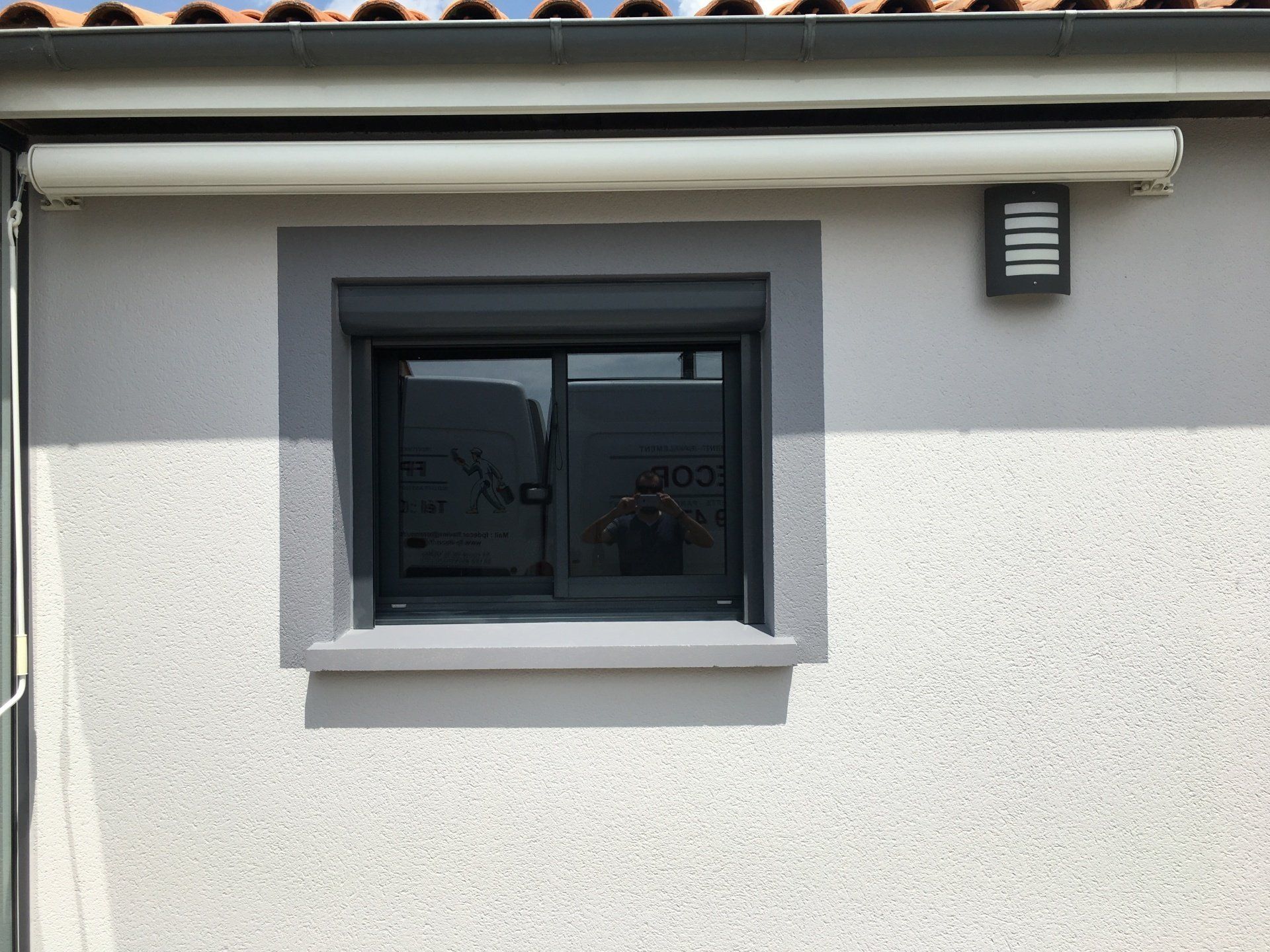 Une fenêtre grise encadrée, encastrée dans un mur blanc texturé, avec un luminaire extérieur noir et un auvent au-dessus.