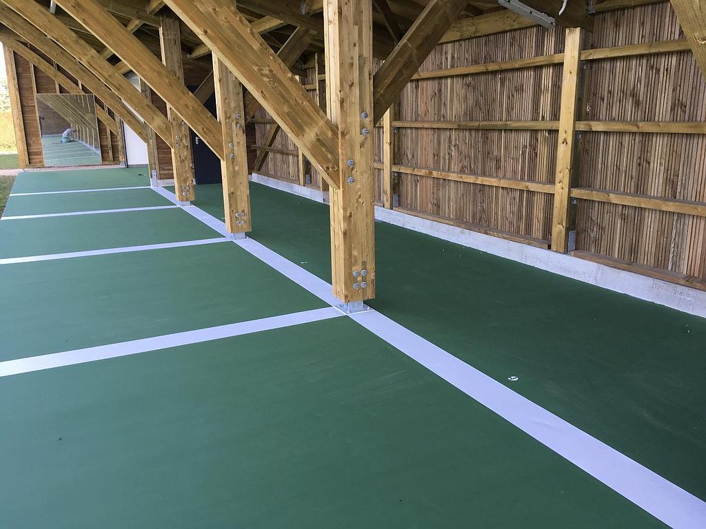 Une structure extérieure en bois avec des poutres apparentes au-dessus d'un sol vert peint de lignes blanches représentant des places de parking.