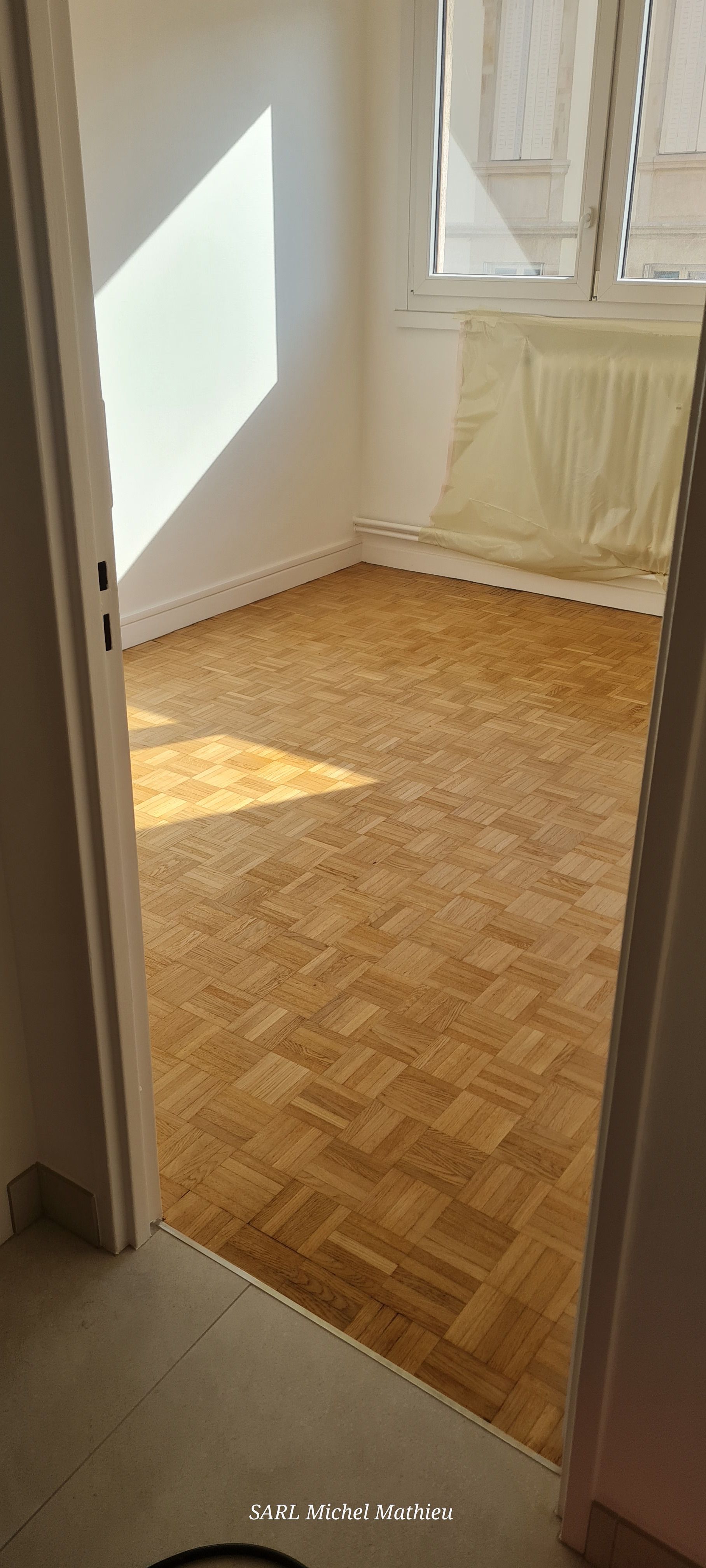 Une pièce avec un parquet, des murs blancs et une fenêtre laissant entrer la lumière du soleil