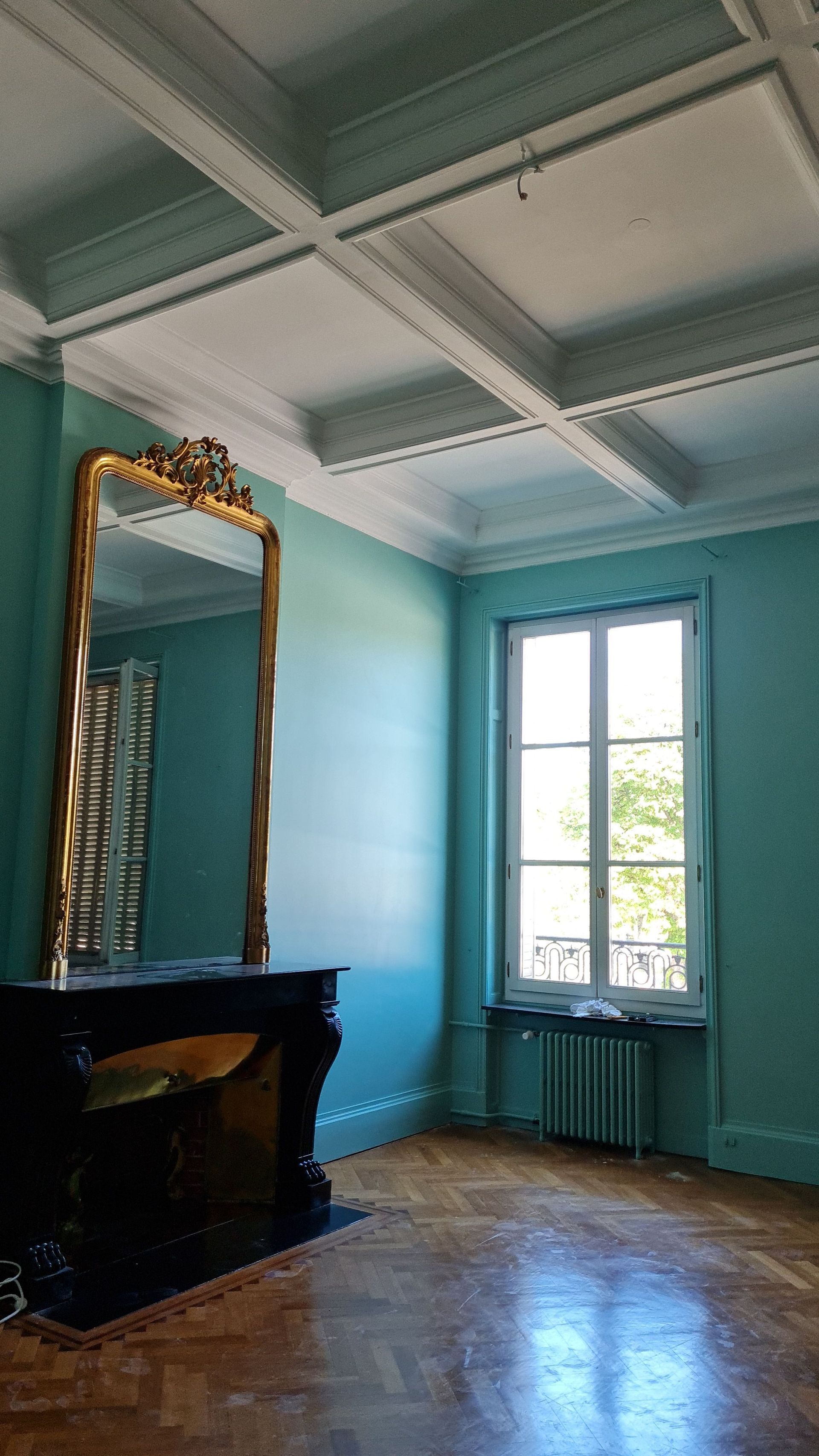 Pièce vide aux murs turquoise, parquet, miroir orné et fenêtre