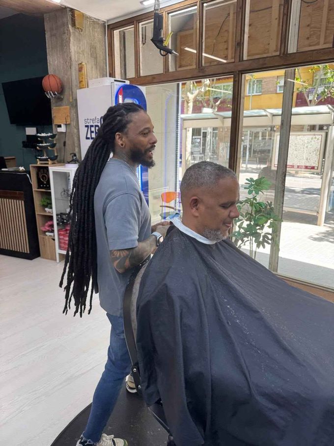 Barbero con rastas cortando el pelo a un cliente en una barbería con una soleada vista exterior.