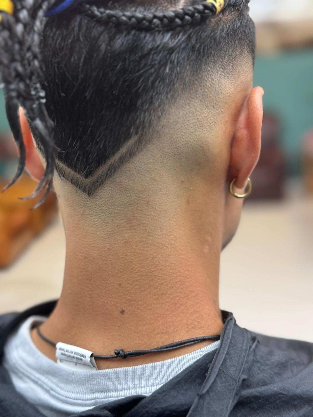 Nuca de una persona con un corte de pelo rapado y descolorido que presenta un diseño en forma de V. Se ven cabello trenzado y un pendiente de oro.