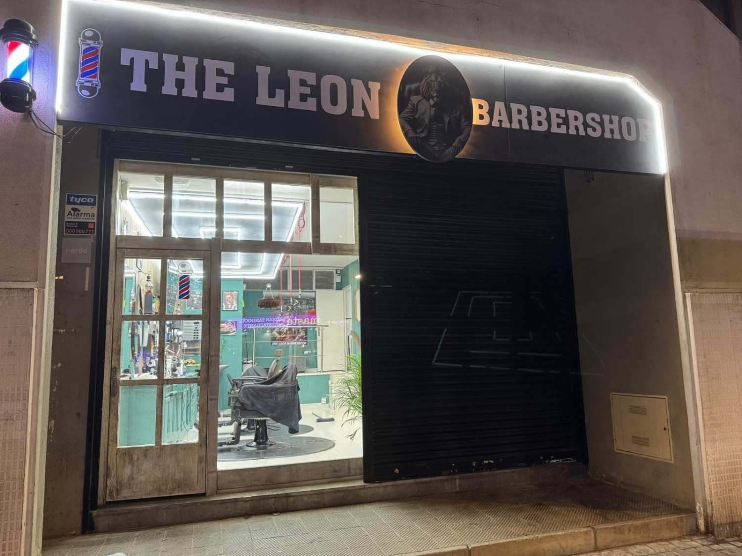 Entrada de una barbería de noche con la puerta abierta y la persiana parcialmente cerrada. Letrero de neón: «La Barbería León».