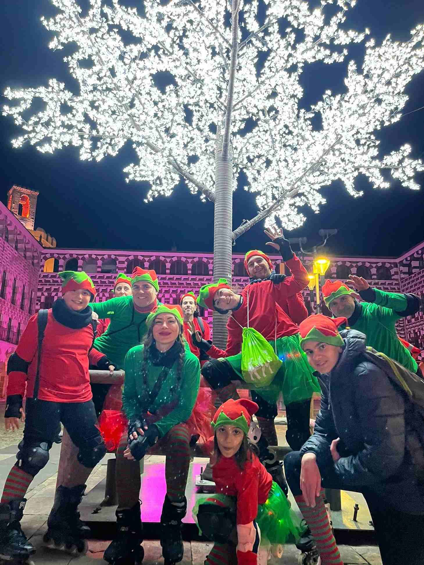Grupo de personas disfrazadas de elfos posando al aire libre por la noche frente a un árbol de luz blanca.
