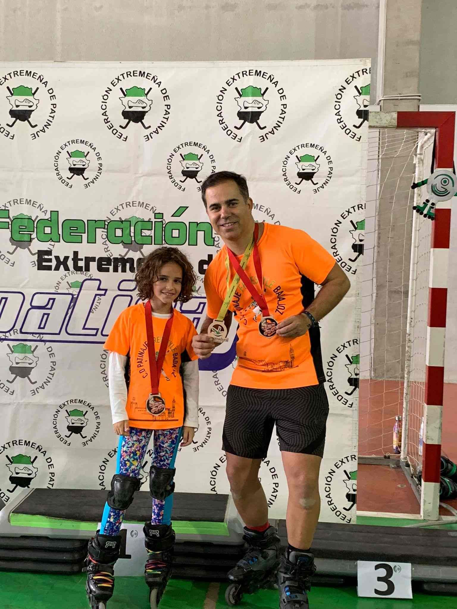 Padre e hija, ambos con camisetas y medallas naranjas, posan en un podio después de una prueba de patinaje.