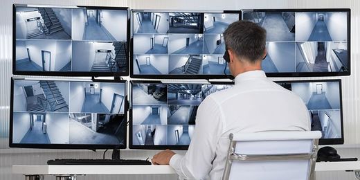 Une personne surveille les flux vidéo de plusieurs caméras de sécurité sur un mur d'écrans dans une salle de contrôle.