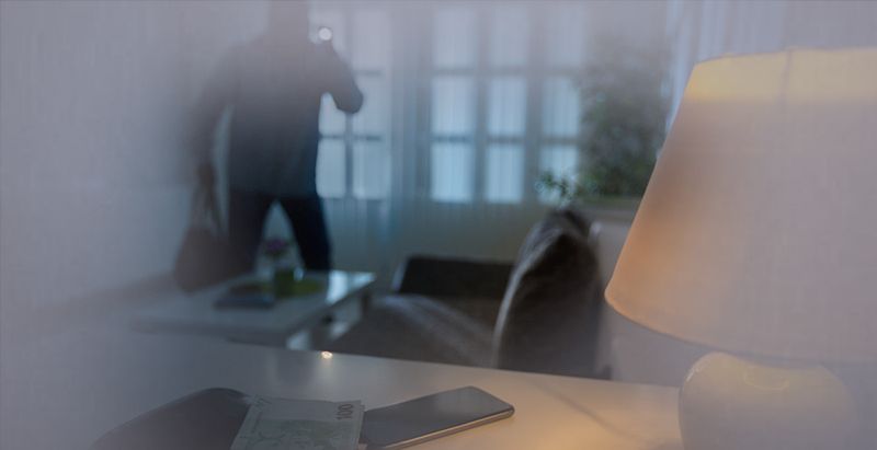 Une pièce sombre aperçue par une fenêtre, avec une silhouette floue près d'une fenêtre lumineuse et une lampe sur un bureau.