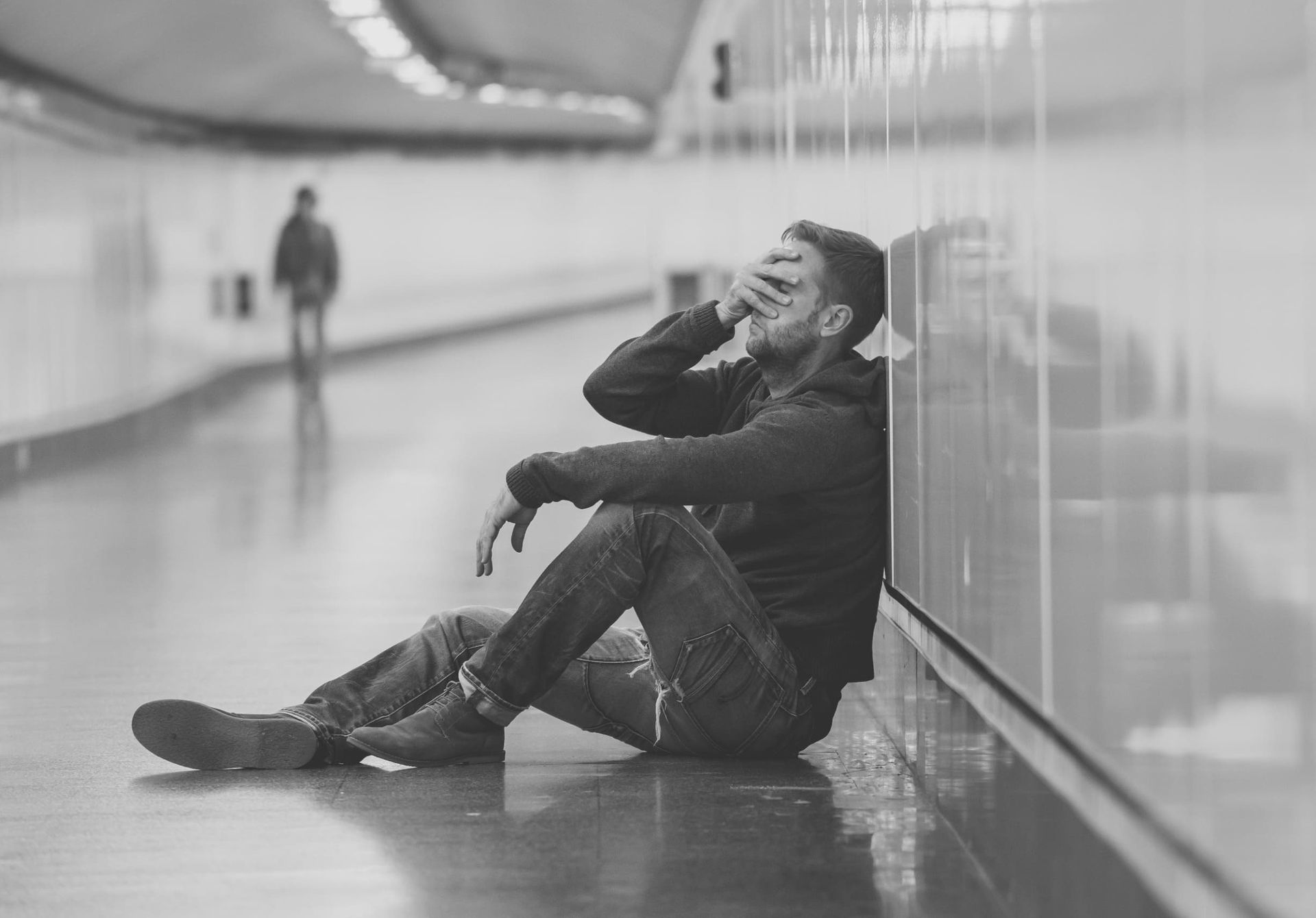 Un hombre está sentado en el suelo apoyado contra una pared cubriéndose la cara.