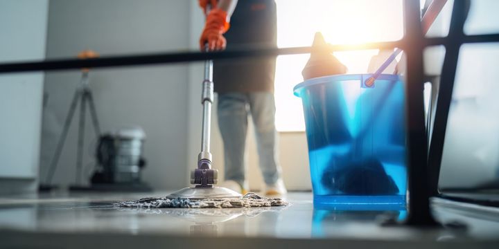 Persona fregando el suelo con fregona y cubo, con guantes naranjas. Luz solar brillante y un trípode al fondo.