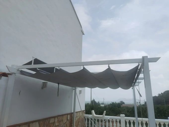 Pérgola blanca con sombra retráctil gris en un balcón, cielo nublado de fondo.