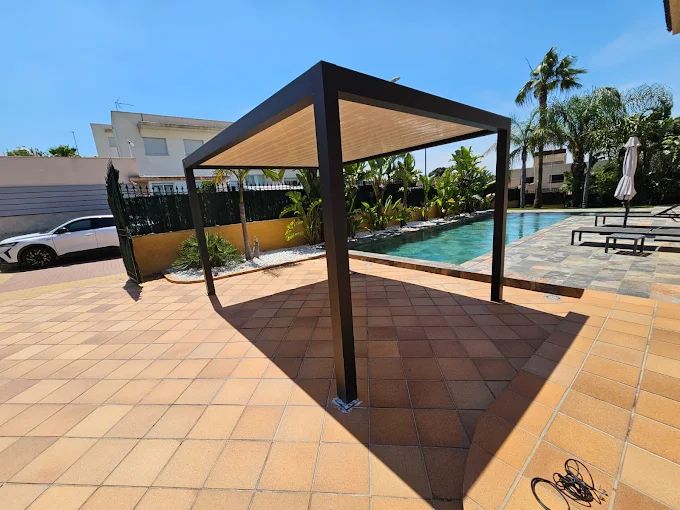 Pérgola que da sombra a un patio de baldosas cerca de una piscina, con un automóvil y una casa visibles.