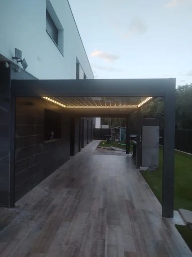 Una cochera moderna con iluminación cálida. Estructura gris oscuro, suelo de madera y una casa blanca al fondo.