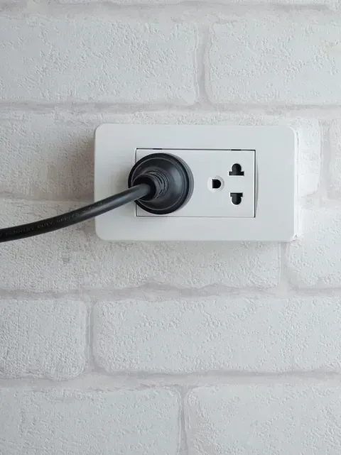 Toma de corriente eléctrica de color blanco con un enchufe negro insertado, montada en una pared de ladrillo blanco.