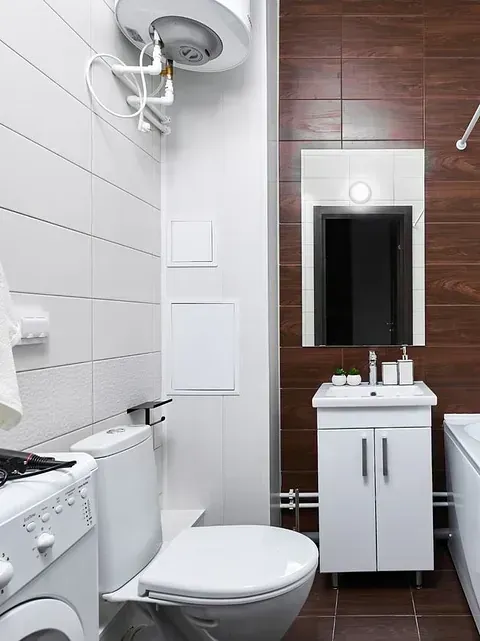 Pequeño baño moderno con paredes de azulejos blancos, inodoro, tocador y lavadora. Un calentador de agua cuelga sobre el inodoro.