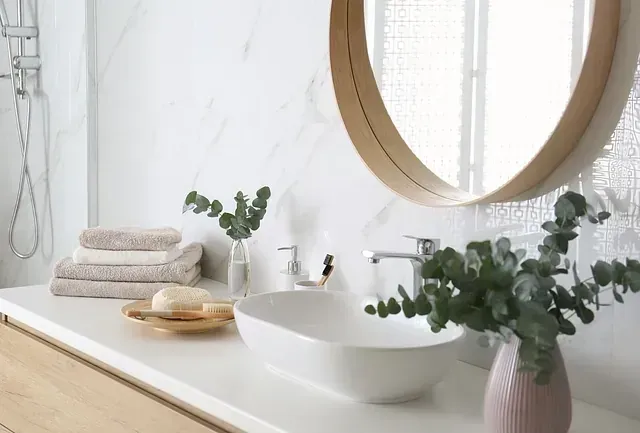 Baño moderno con encimera blanca, lavabo ovalado y ramas de eucalipto. Toallas beige y un espejo redondo completan el conjunto.