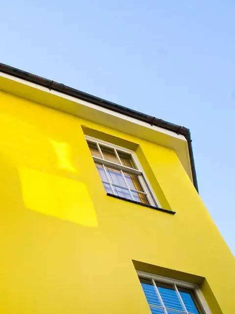 Edificio amarillo con dos ventanas con marco blanco contra un cielo azul claro.
