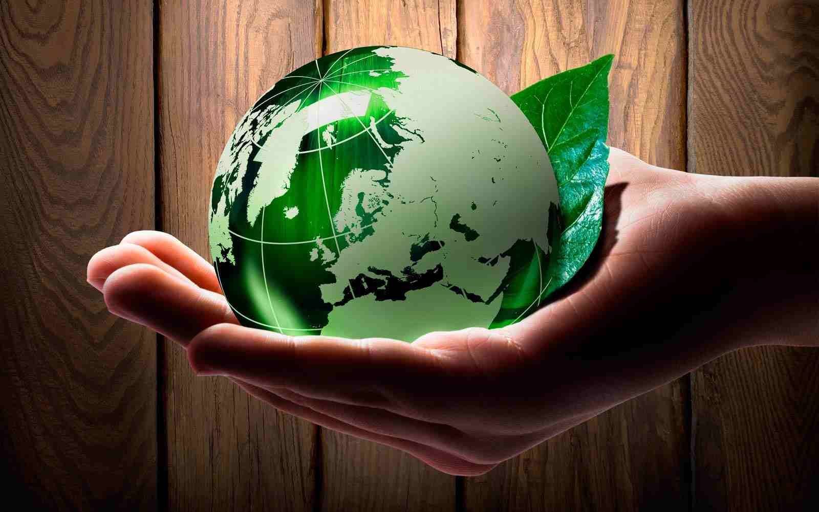 Una mano sostiene un globo terráqueo verde translúcido junto a una hoja verde sobre un fondo rústico de madera.