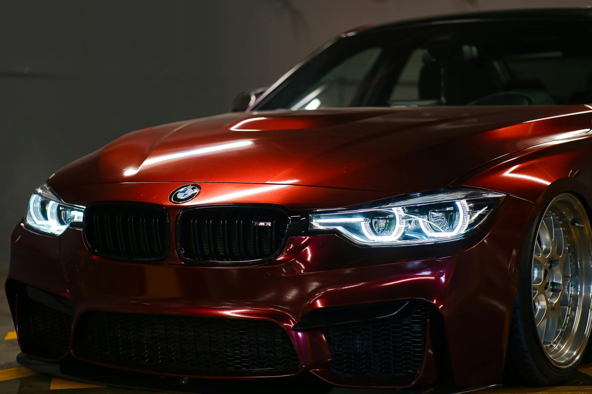 Un deportivo BMW M3 de color rojo metalizado brillante, con faros LED iluminados, estacionado en un garaje con poca luz.