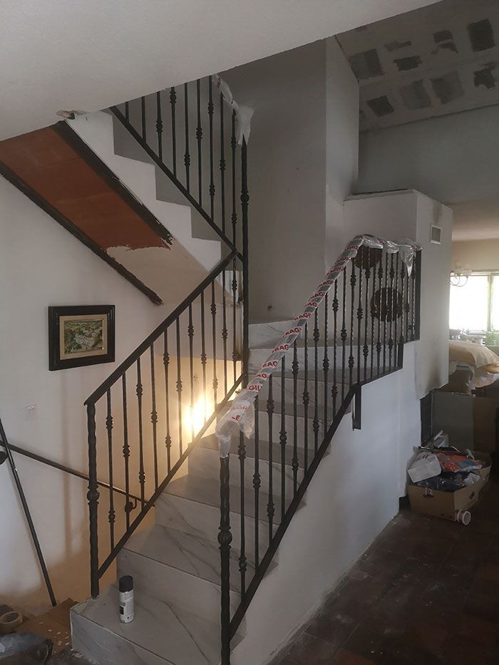 Escalera con escalones de mármol, barandilla de hierro forjado negro, paredes blancas y estantería de madera.