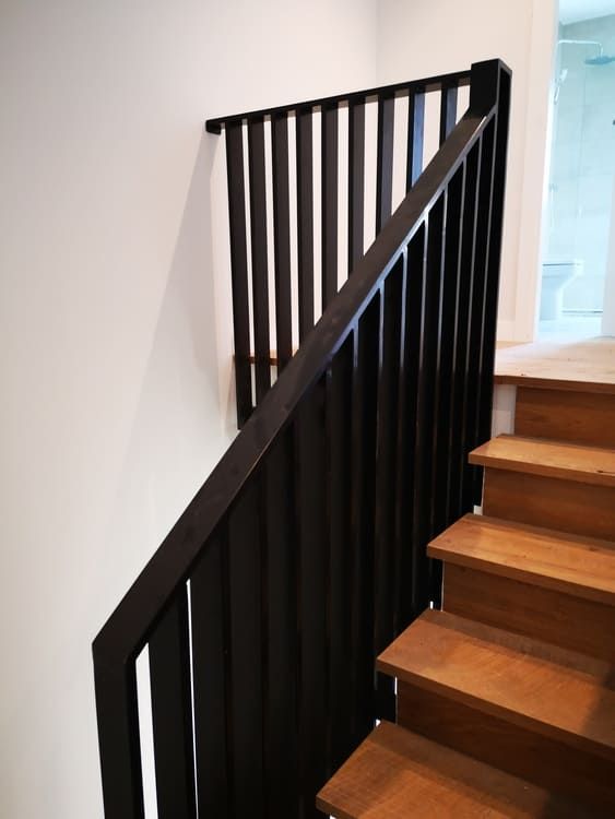 Escalera de madera con barandilla vertical negra sobre una pared blanca.