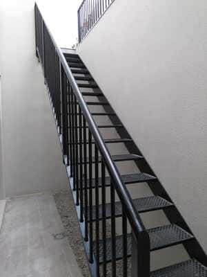 Escalera de metal negro contra una pared de color claro con una barandilla negra que conduce hacia arriba.