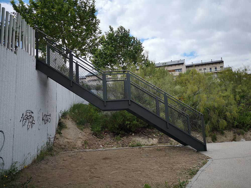 Escalera de metal con pasamanos a lo largo de una pared de hormigón, que conduce a una ladera con follaje y edificios.