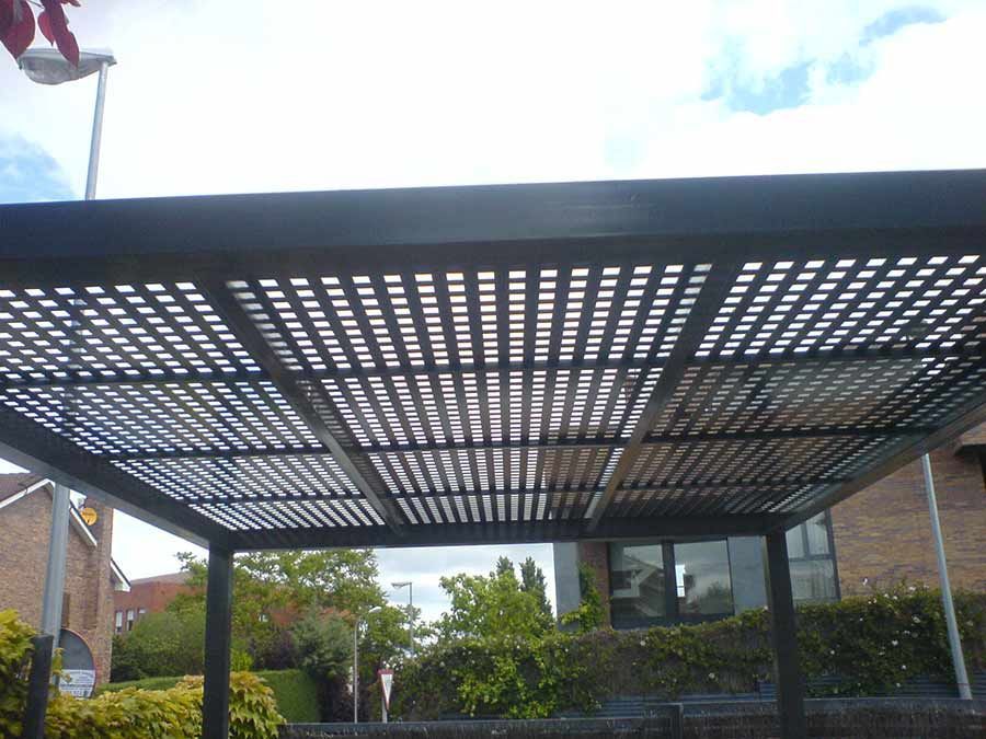 Pérgola de celosía de color gris oscuro sobre un espacio exterior con árboles circundantes y un edificio de ladrillo.