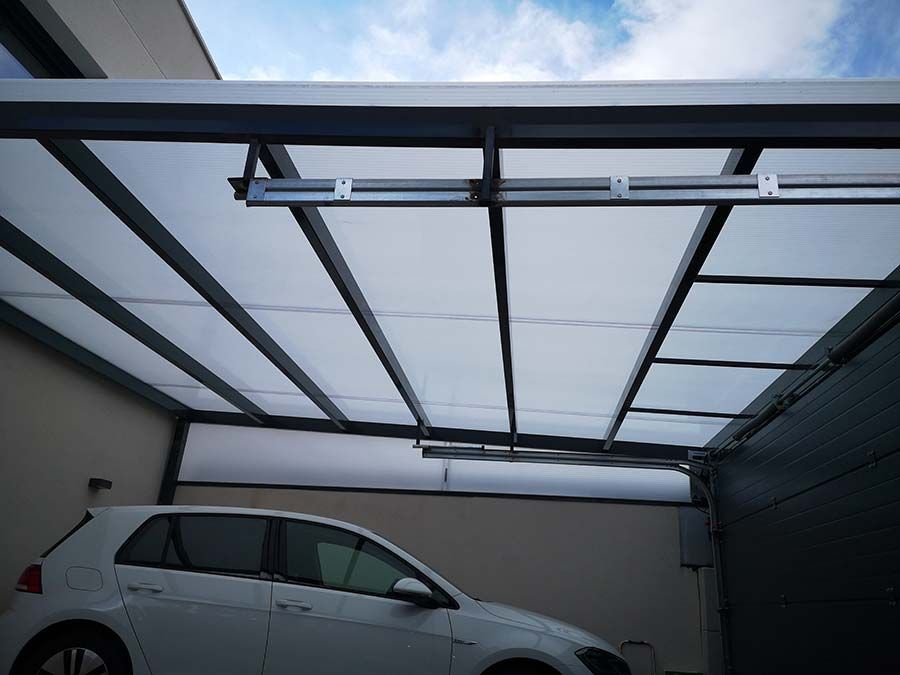 Coche blanco aparcado bajo una cochera con techo transparente. Chasis metálico negro visible. Cielo azul.