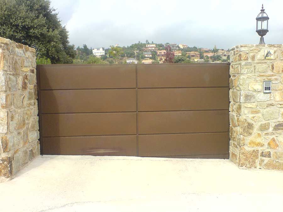Puerta de entrada de metal marrón entre pilares de piedra, colocada en hormigón, con edificios al fondo.