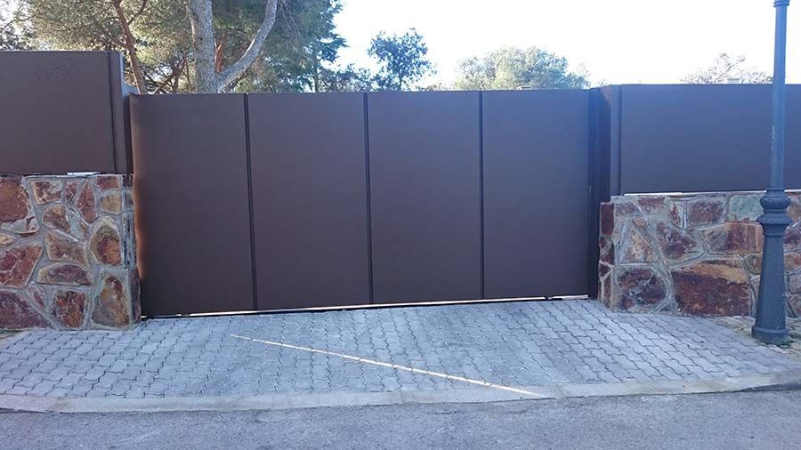 Puerta de metal marrón con pilares de piedra y camino de entrada de ladrillo.